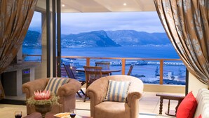 A room with a view of a body of water and mountains, featuring a sofa, armchair, and dining table with a vase of flowers and a wine glass. - Mariner Guesthouse (Cape Town)