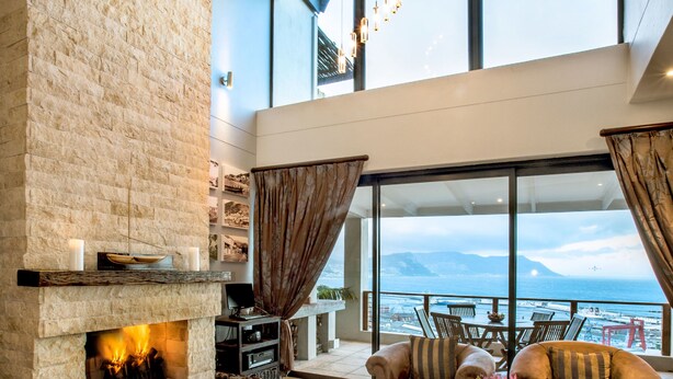A spacious living room with a stone fireplace, comfortable seating, a coffee table with magazines, and a large window offering a view of the sea. - Mariner Guesthouse (Cape Town)