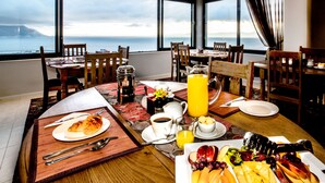 A breakfast setting with a view of the sea, featuring a wooden table, chairs, and a variety of breakfast items including fruits, pastries, and beverages. - Mariner Guesthouse (Cape Town)