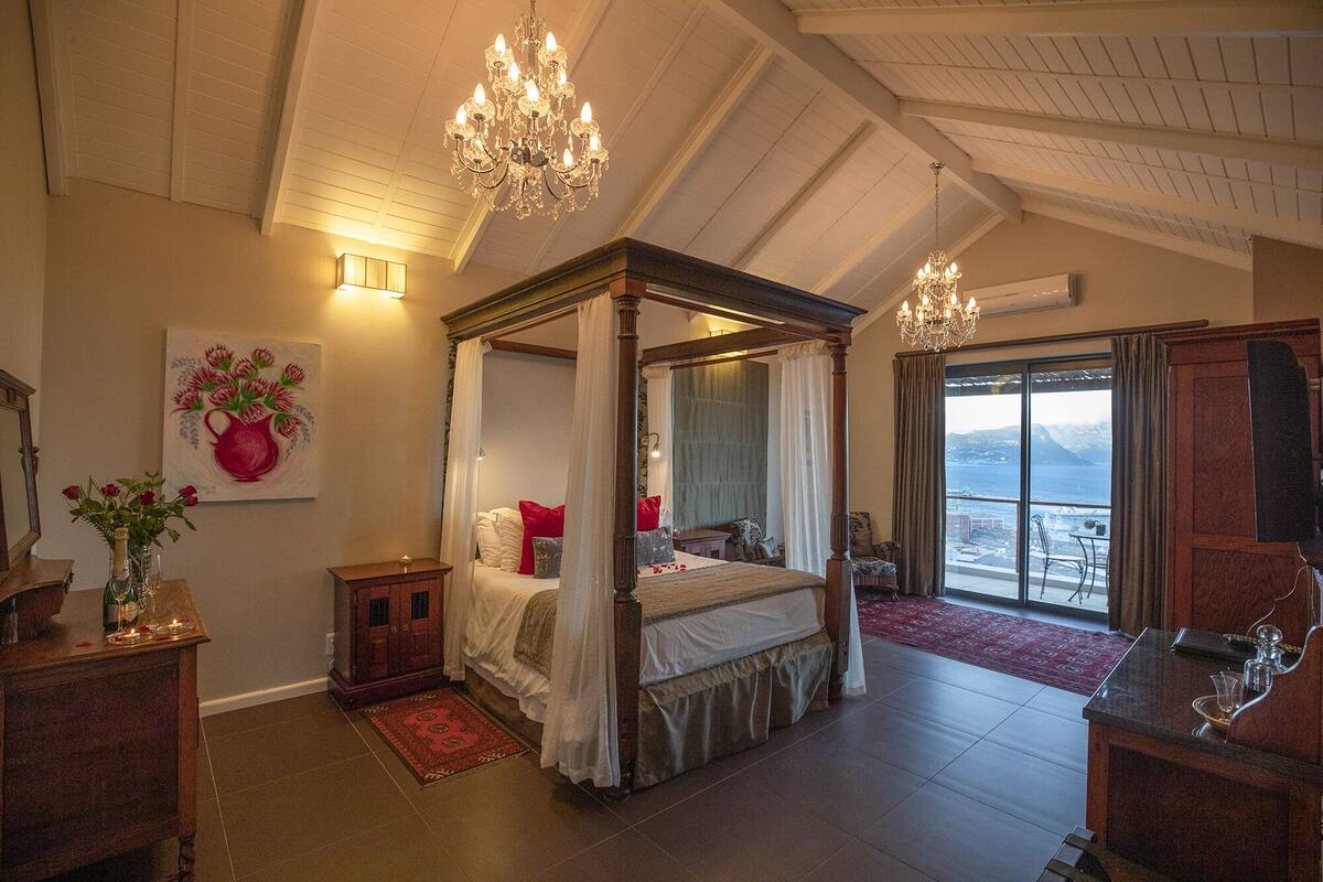Uma cama com dossel e cortina, um lustre, uma pintura de flores e vista para o mar por uma grande janela.