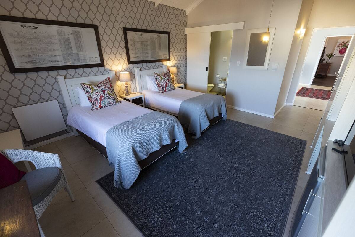 Quarto de hotel com duas camas, um tapete escuro estampado e uma cadeira branca.