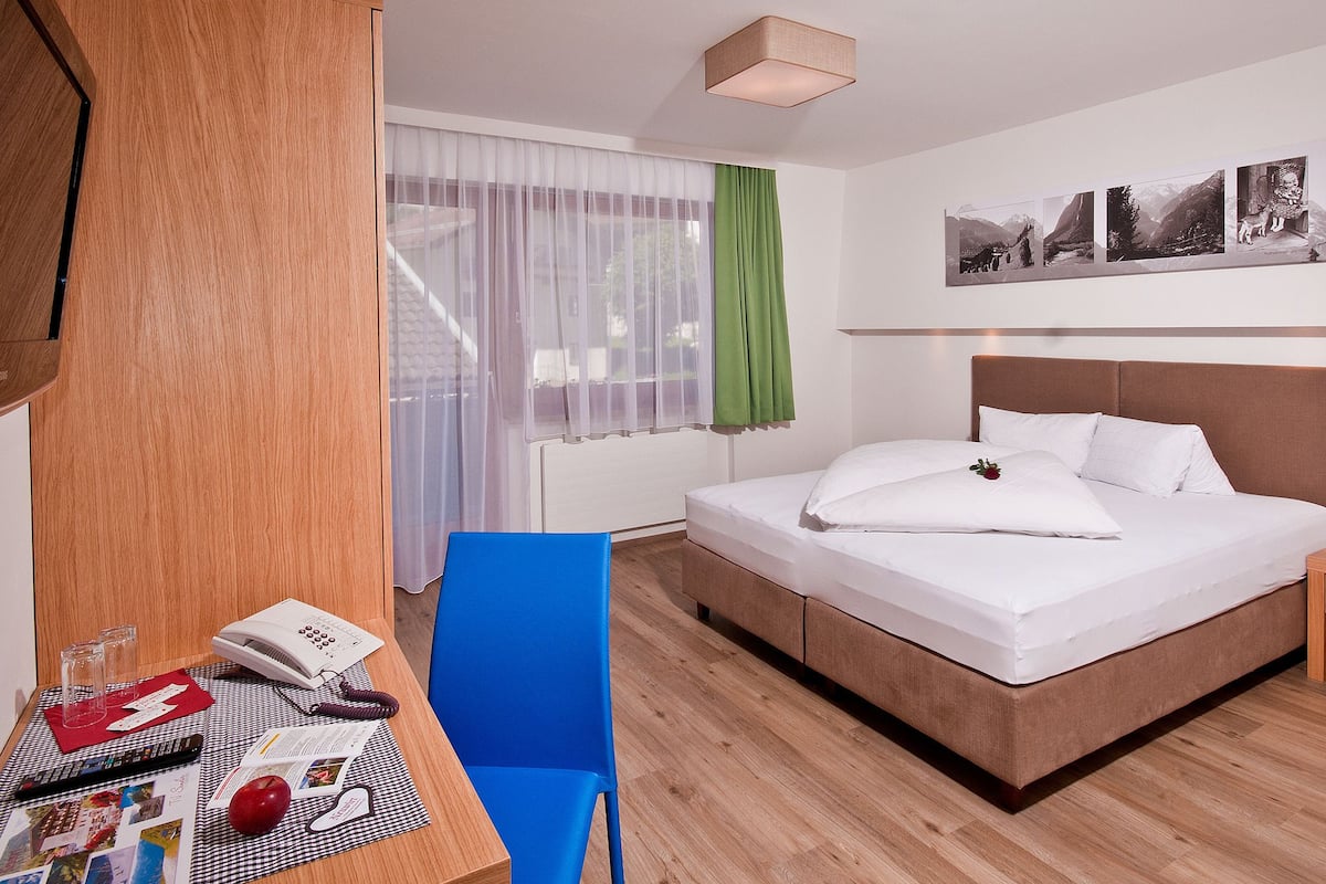 A hotel room with a bed, a desk with a chair, a telephone, and a window with sheer curtains.