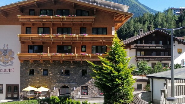 Un hotel amb façana de pedra, balcons amb flors i un paisatge de muntanya al fons.