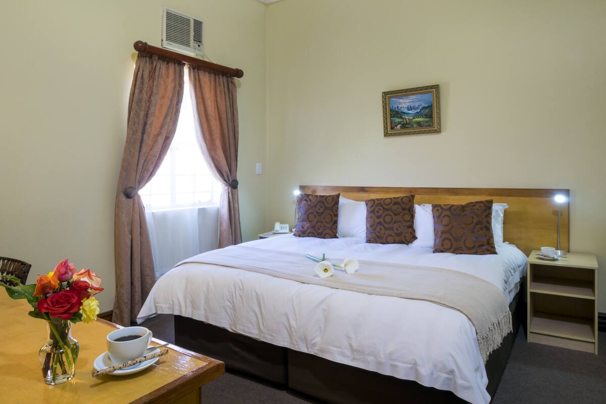 Una habitación de hotel con cama, una mesita de noche con una taza de café, un florero con flores y un cuadro en la pared.