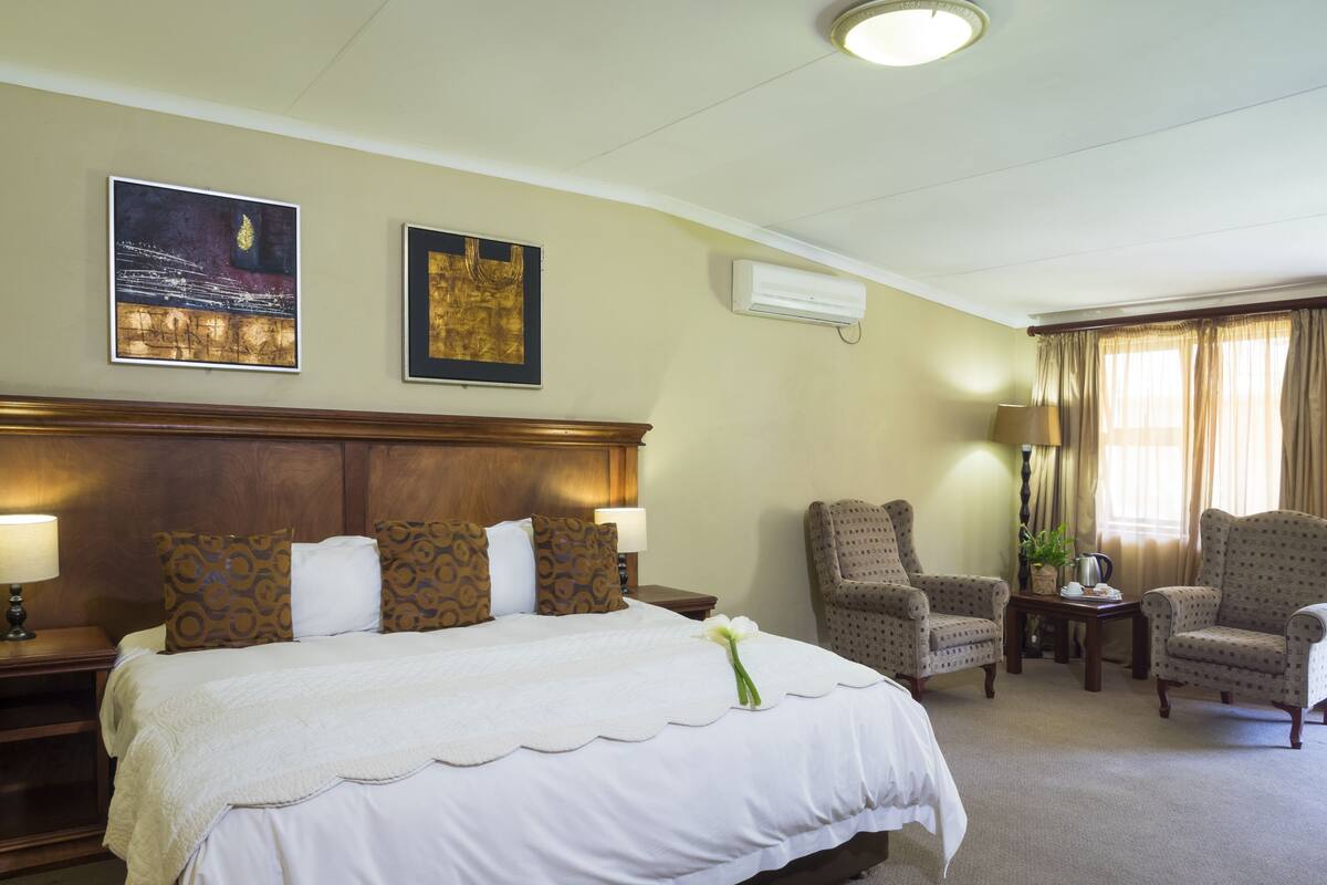 Una habitación de hotel con una cama grande, dos sillones, una mesita con lámpara y un ventanal con cortinas.