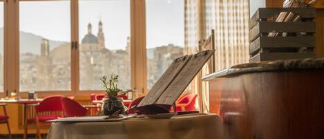 Un restaurante con una mesa dispuesta para dos personas, un atril con el menú y vistas a un paisaje urbano a través de amplias ventanas.