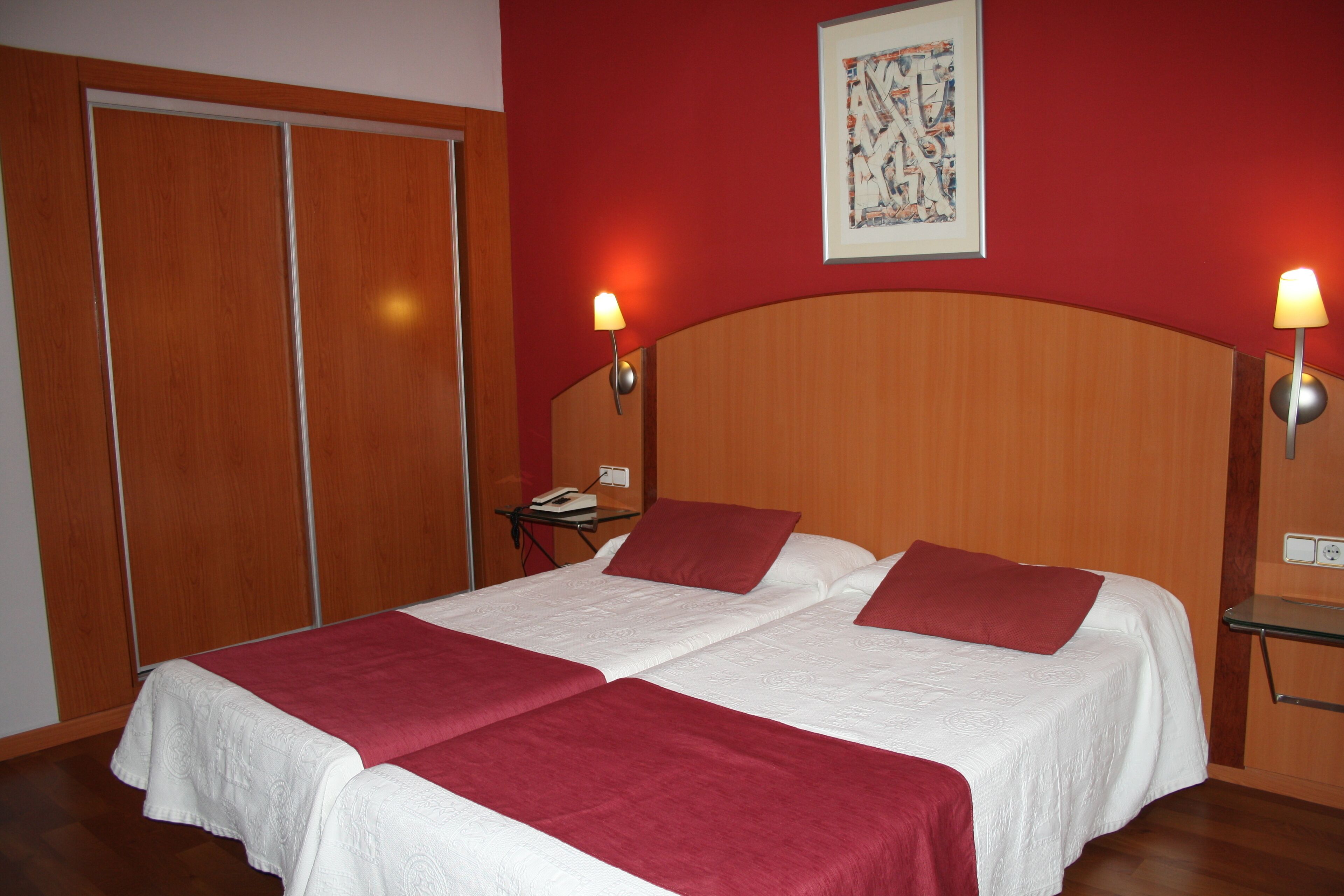 A double bed with white bedding and red throw pillows, a wooden headboard, and two bedside lamps.
