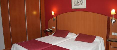 Cama doble con ropa de cama blanca y almohadas rojas decorativas, cabecera de madera y dos lámparas de noche.