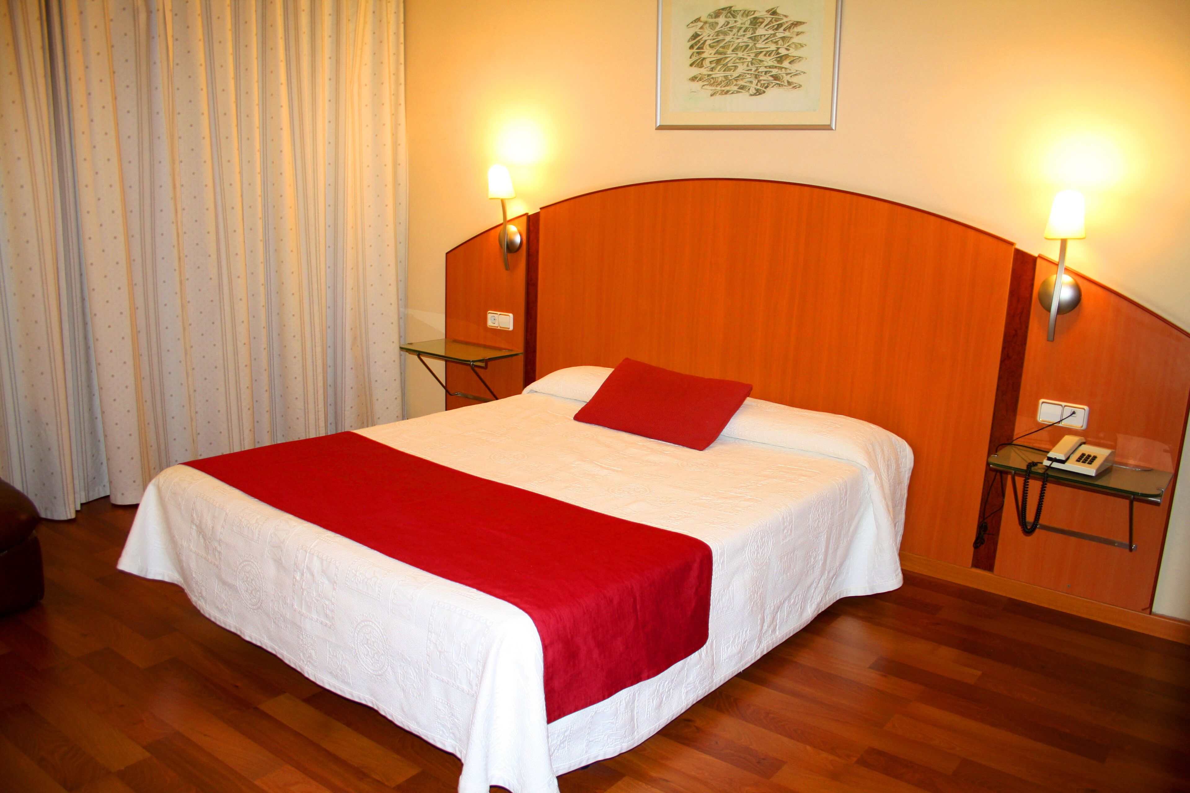 Camera d'albergo con un letto grande, una coperta rossa, una testiera in legno, due lampade da comodino, un piccolo tavolo con il telefono e una tenda alla finestra.