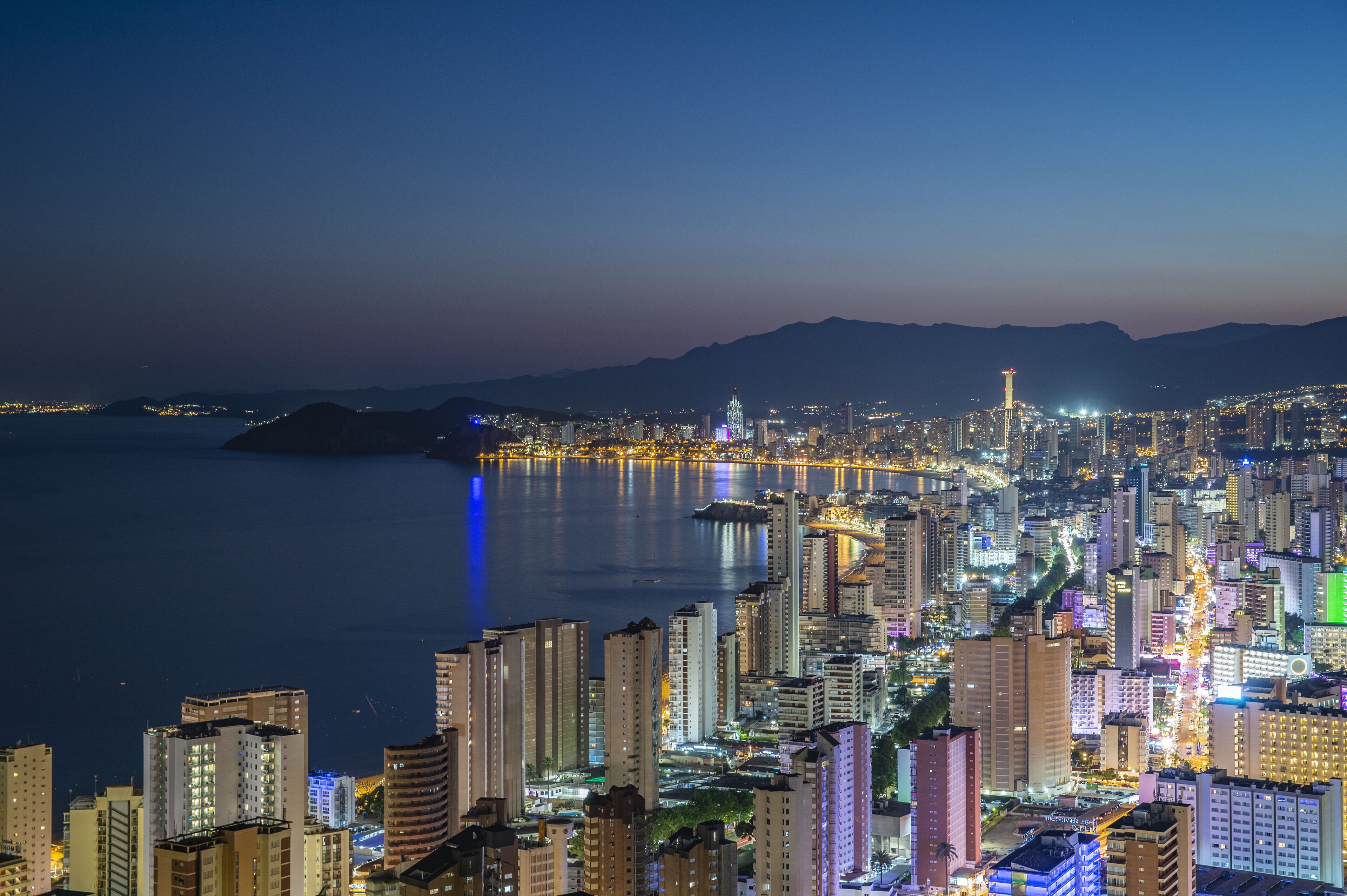 A cityscape at night with high-rise buildings, a waterfront, and a mountain range in the background.