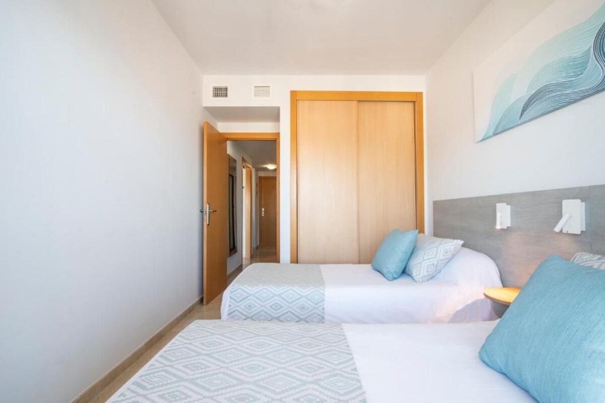 A hotel room with two beds, a wooden door, a gray headboard, and a wall-mounted light fixture.