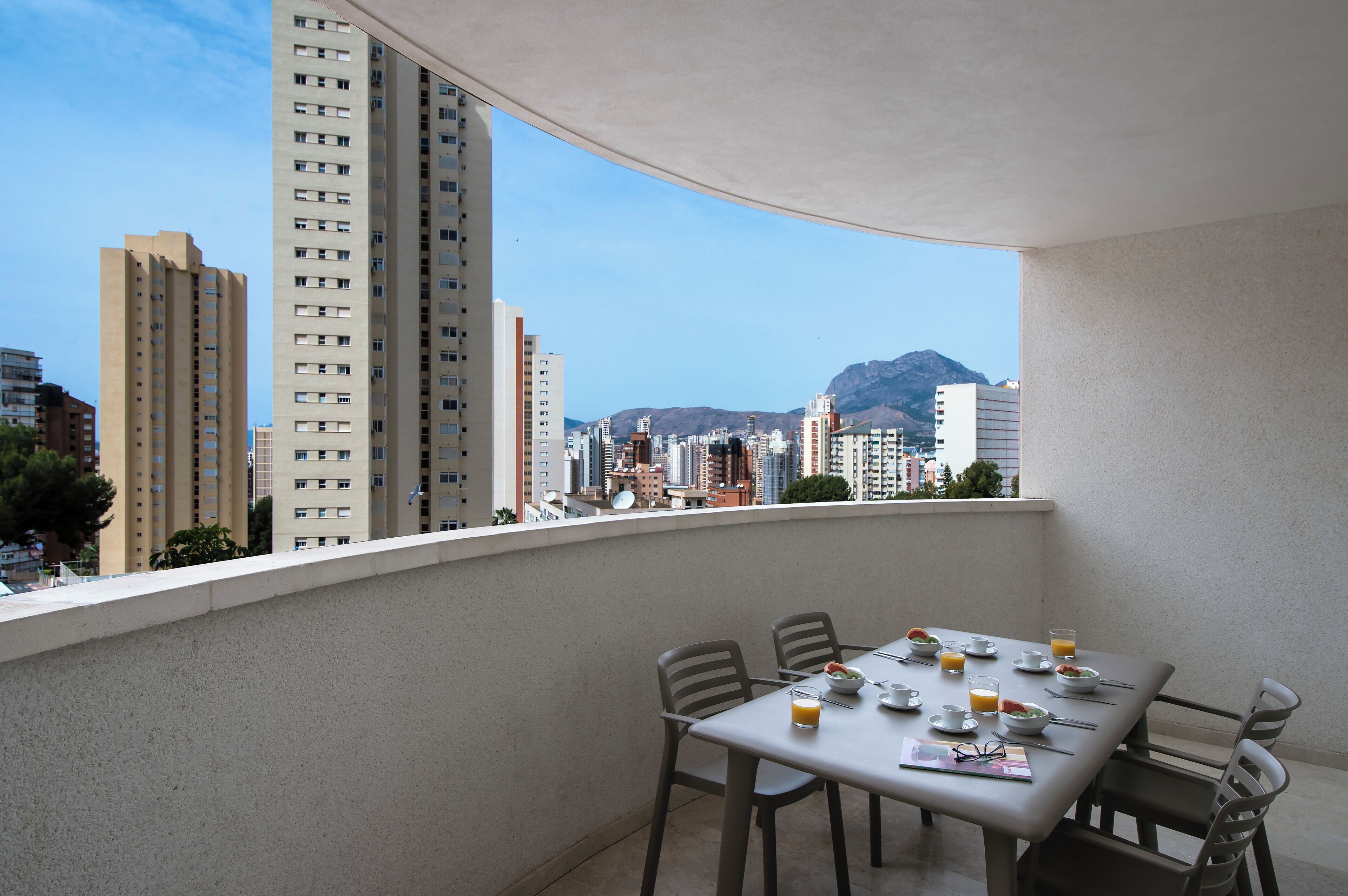 Un balcone con un tavolo apparecchiato per la colazione, con vista su un paesaggio urbano caratterizzato da alti edifici e montagne.