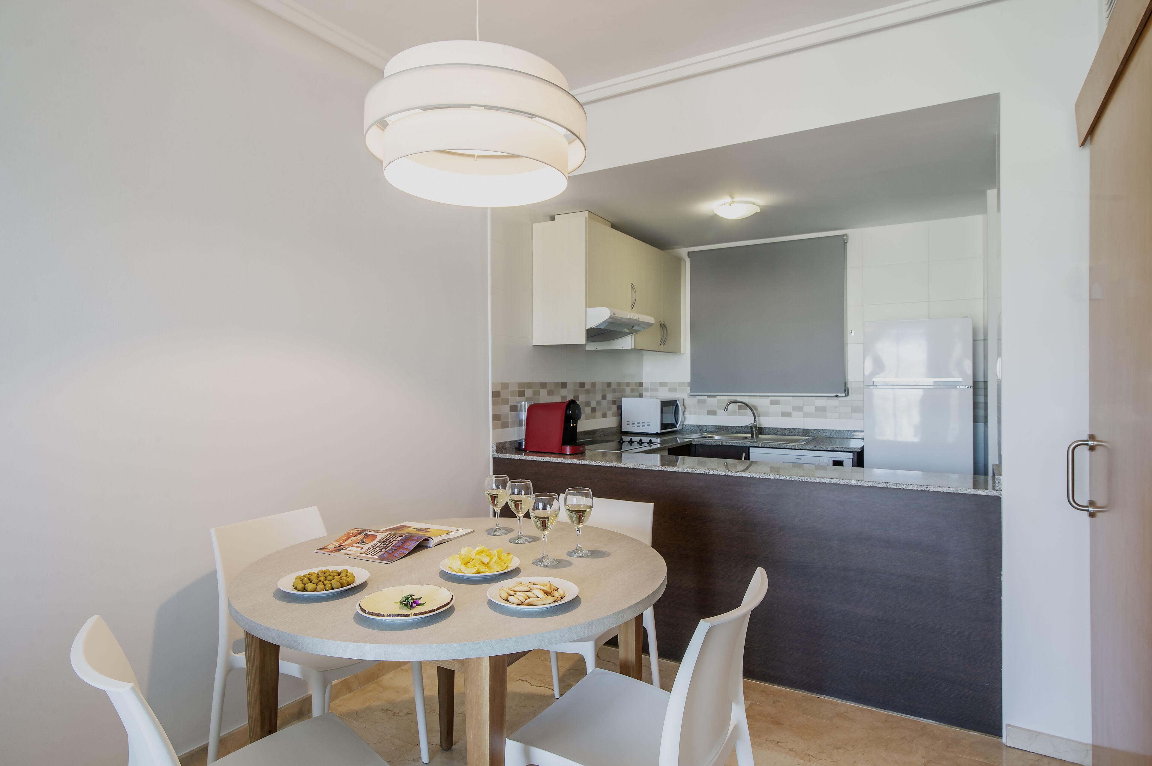 Una cocina compacta con electrodomésticos modernos, una zona de comedor con una mesa redonda y sillas, y una lámpara colgante.
