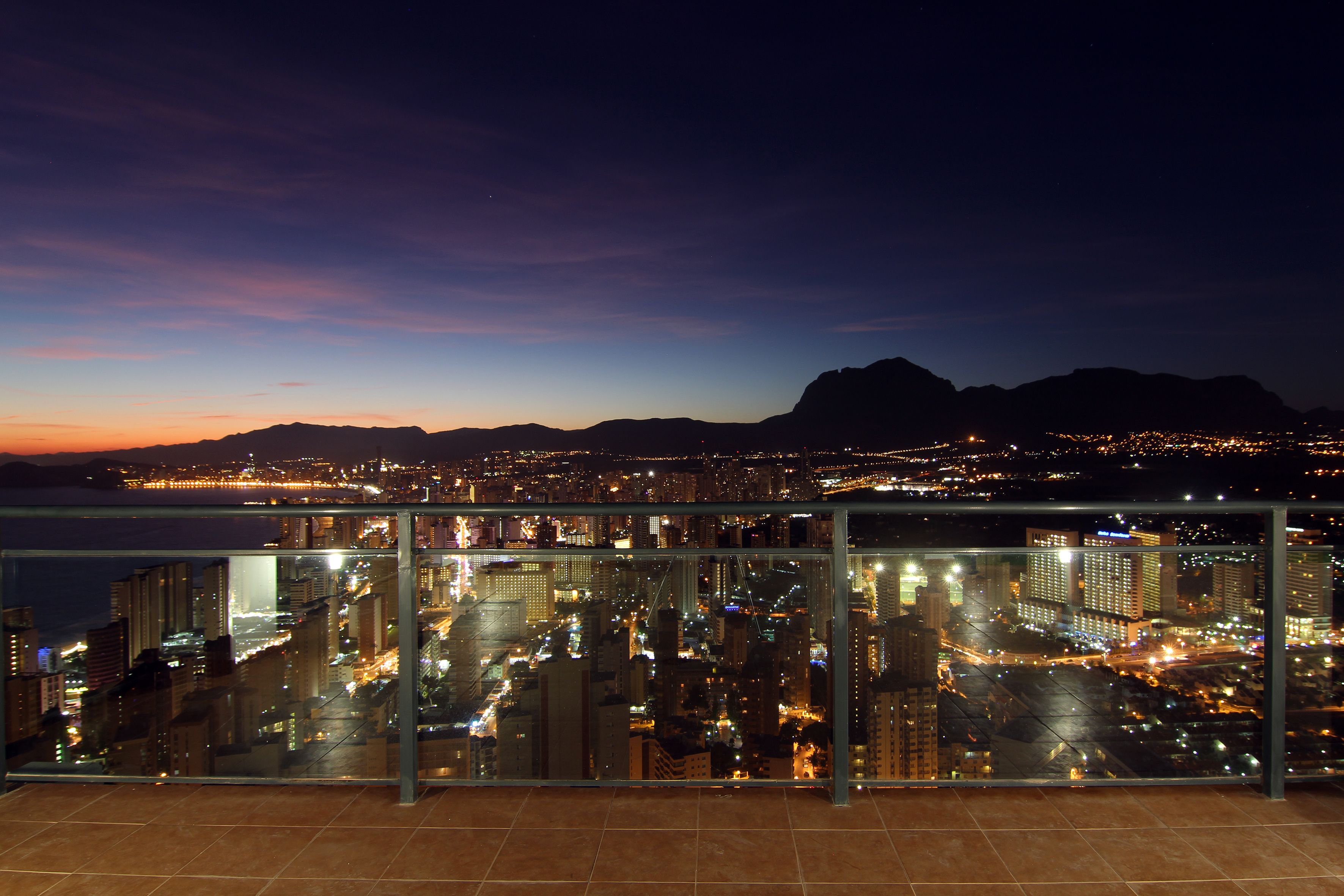 A cityscape at dusk with a mountain in the background and a view from a balcony.