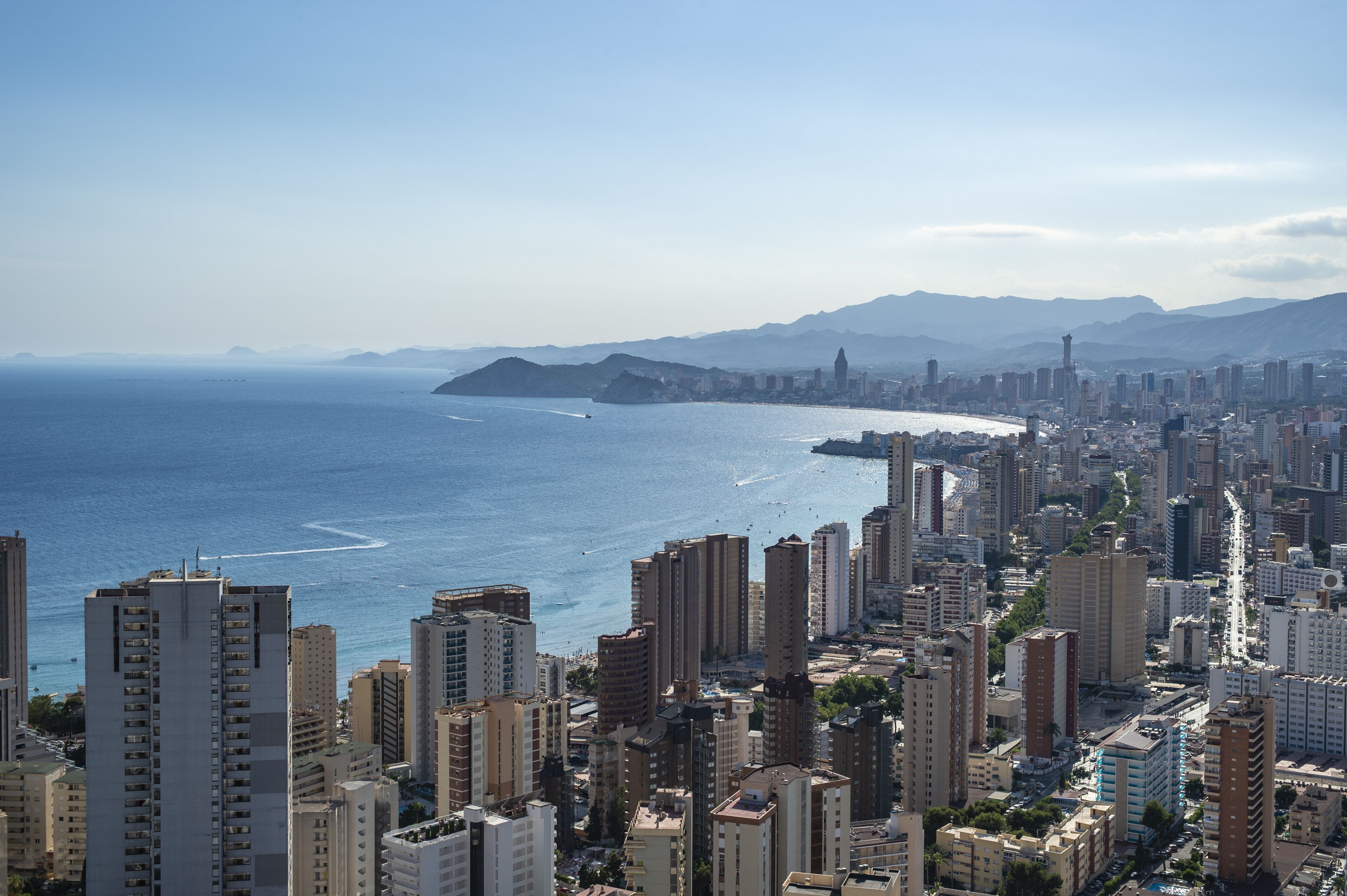 A coastal cityscape with high-rise buildings, a clear view of the sea, and distant mountains.