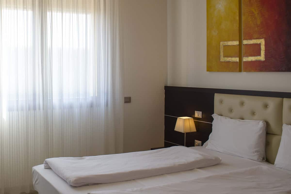 Ein Hotelzimmer mit zwei Betten, einem großen Fenster mit leichtem Vorhang, einem Nachttisch mit Lampe und einem an der Wand hängenden Kunstwerk.