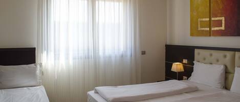Habitación de hotel con dos camas, un ventanal con cortinas translúcidas, una mesita de noche con lámpara y una obra de arte colgada en la pared.