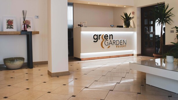 A reception area with a 'Green Garden Resort' sign, a reception desk, a glass-top table, and potted plants. - Green Garden Resort (Mestre)