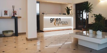 Un'area reception con il cartello 'Green Garden Resort', una scrivania e tavolini in vetro, con piante in vaso.