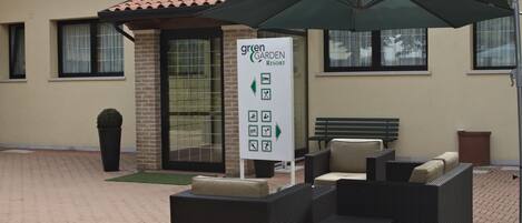 Un área de estar al aire libre con mobiliario de mimbre, un paraguas verde, un letrero que dice 'Green Garden Resort' y un muro de ladrillo con ventanas.