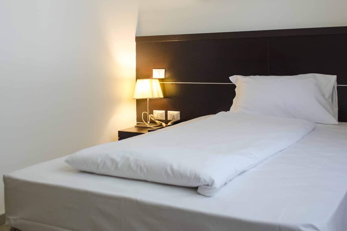 Ein ordentlich gemachtes Bett mit weißen Leinen und Kissen, eine Nachttischlampe und ein Nachttisch mit Telefon und Buch.
