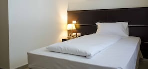 Una cama bien tendida con sábanas y almohadas blancas, una lámpara de noche y una mesita de noche con un teléfono y un libro.