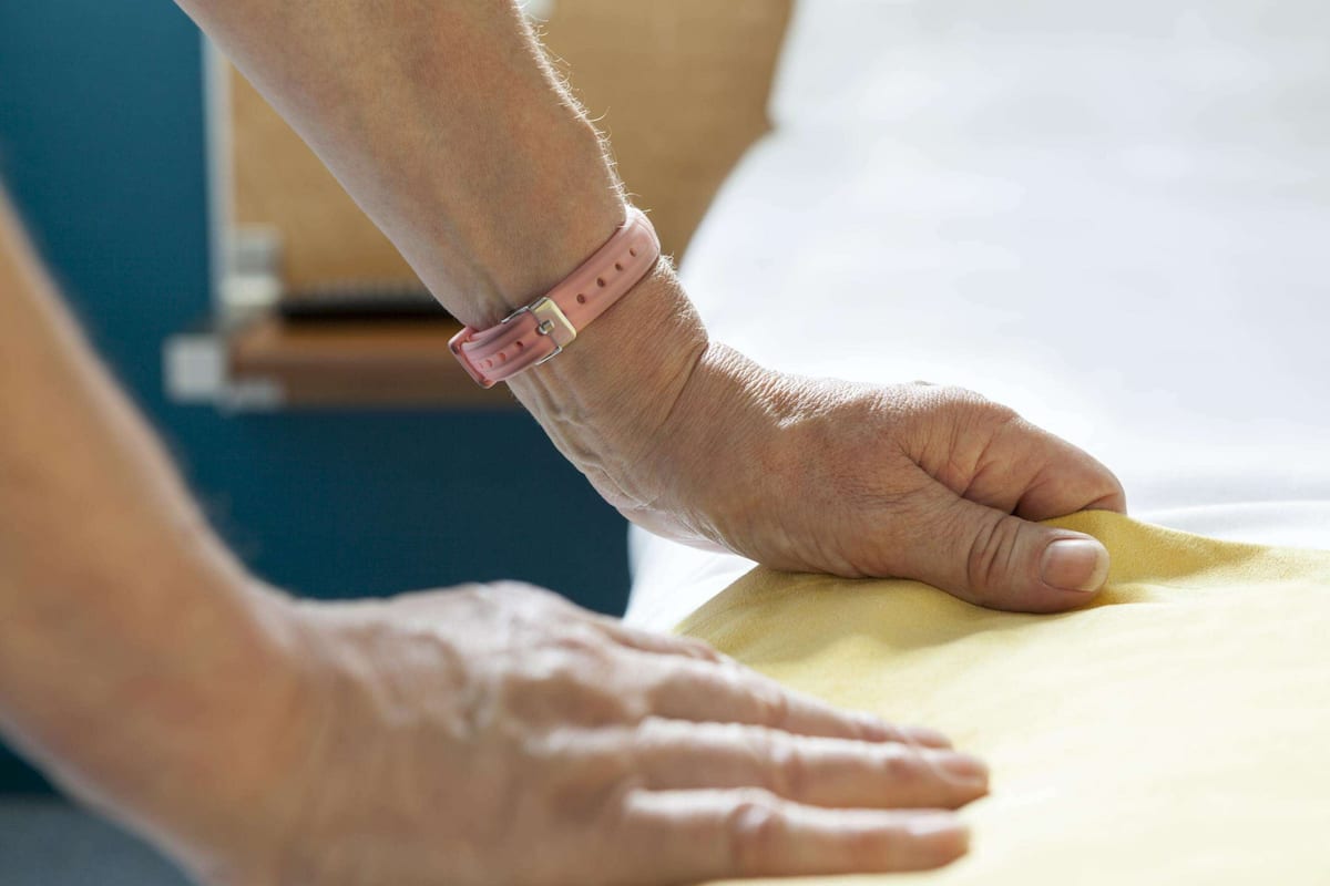 Eine Person mit einer rosafarbenen Armbanduhr befestigt ein gelbes Bettlaken auf einem Bett.