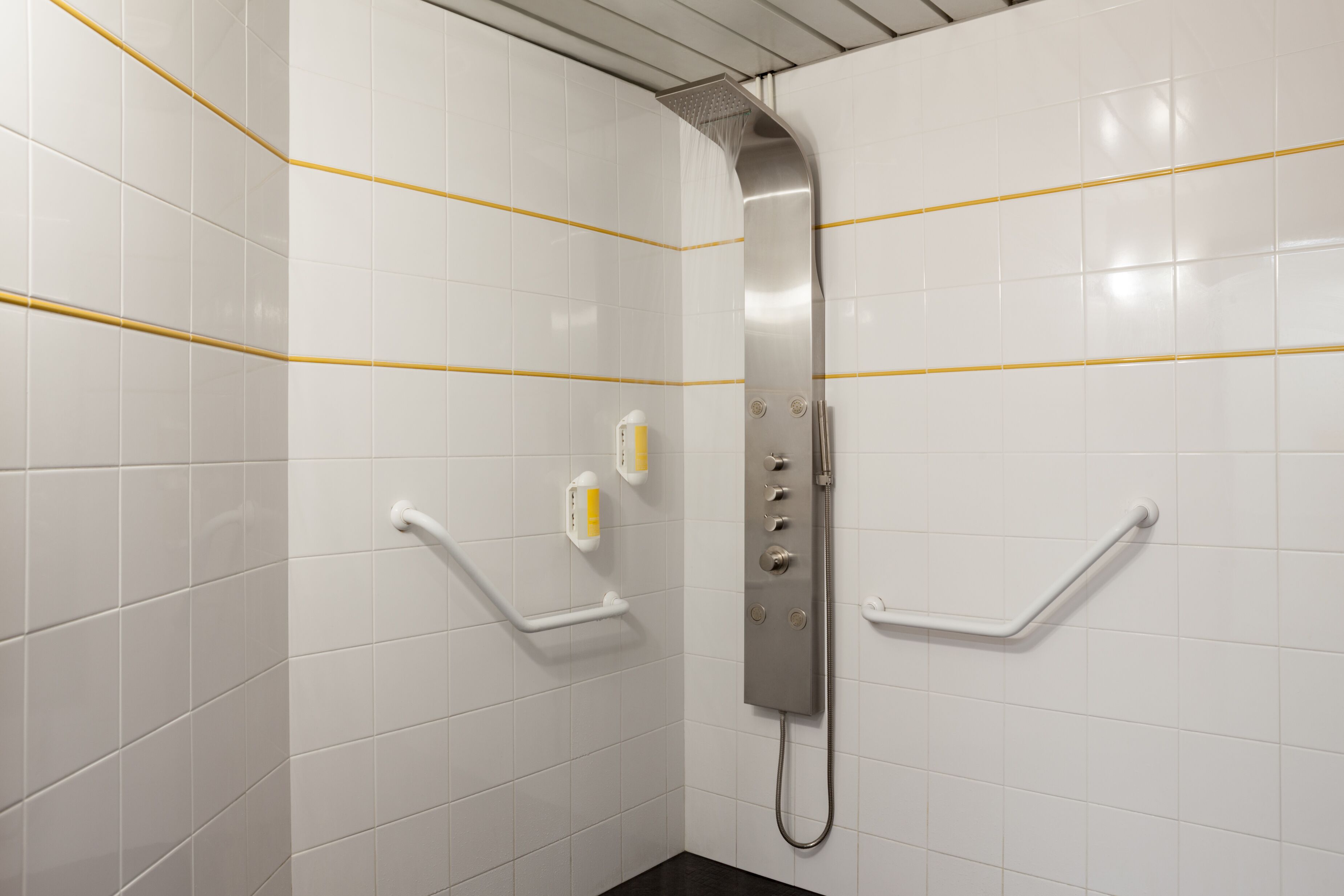 Een badkamer met witte betegelde wanden, een douche met handdouche en handgrepen.