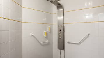 Un baño con paredes de azulejos blancos, una ducha con cabezal de ducha de mano y barras de apoyo.