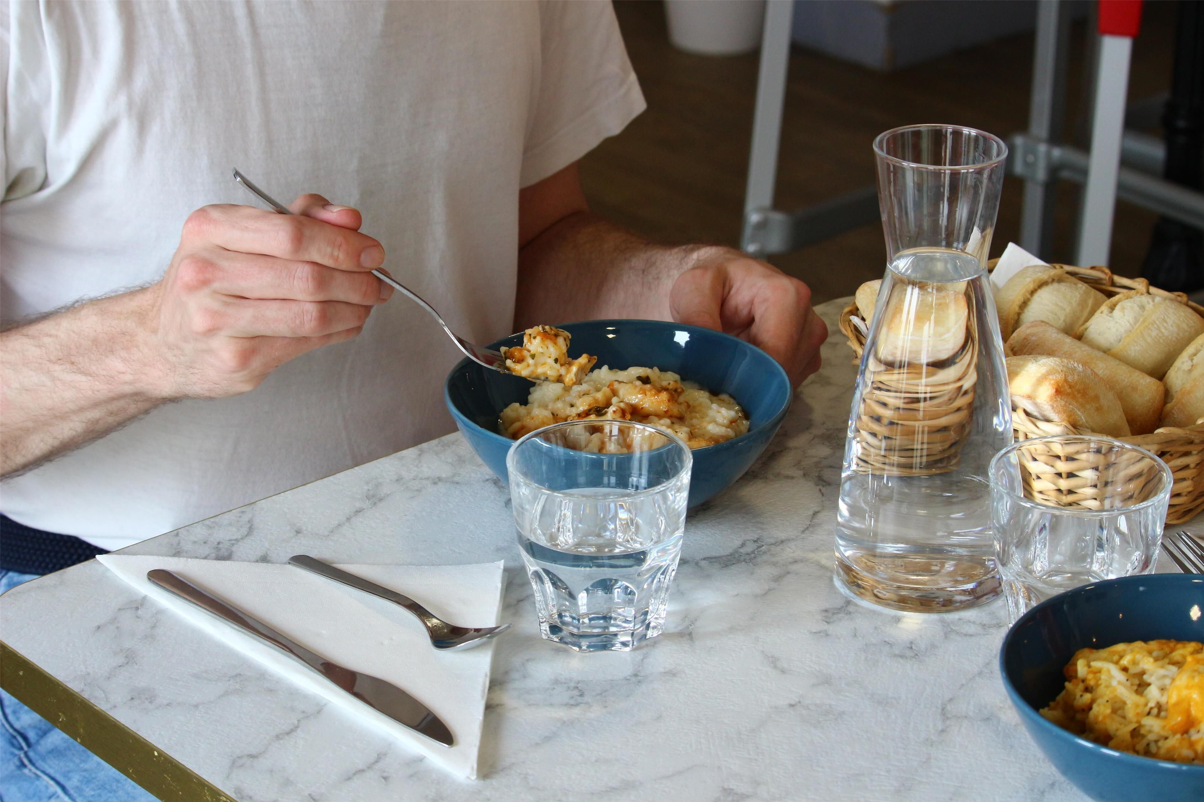 A dining table with a bowl of cereal, a glass of water, a basket of bread, and a clear glass bottle.