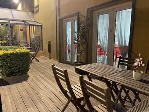 An outdoor patio with wooden tables and chairs, a potted plant, and a glass door leading to the interior.