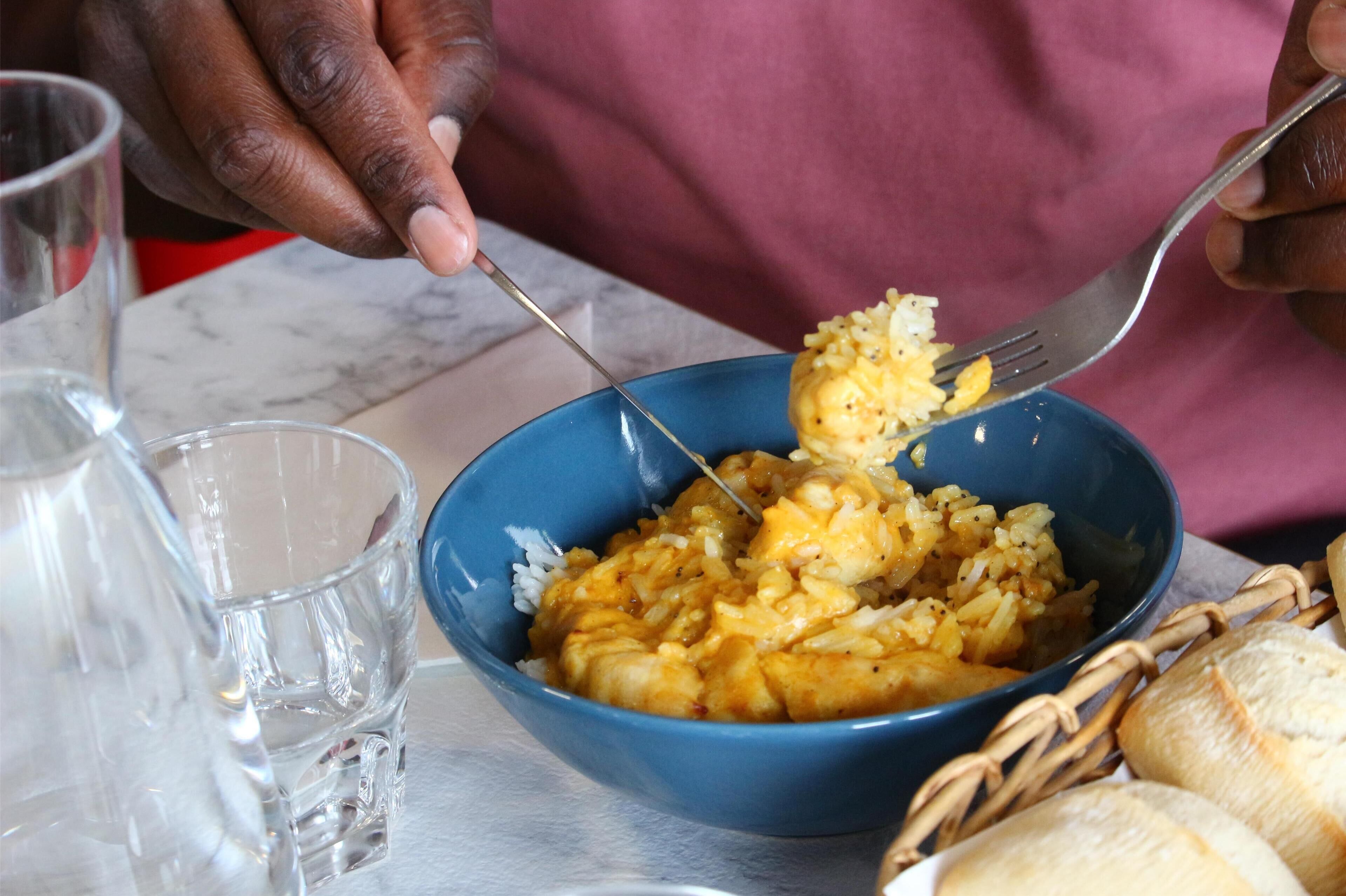 A person eating from a blue bowl with rice and a yellowish dish, possibly curry, with a fork. There is a clear glass and a basket with bread on the table.