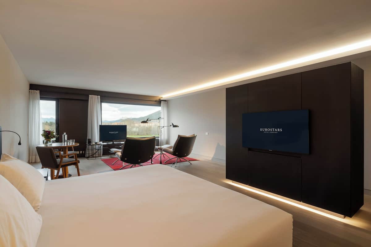 Een moderne hotelkamer met een groot bed, een bureau met twee stoelen, een flatscreen televisie aan de muur en uitzicht op de bergen door het raam.