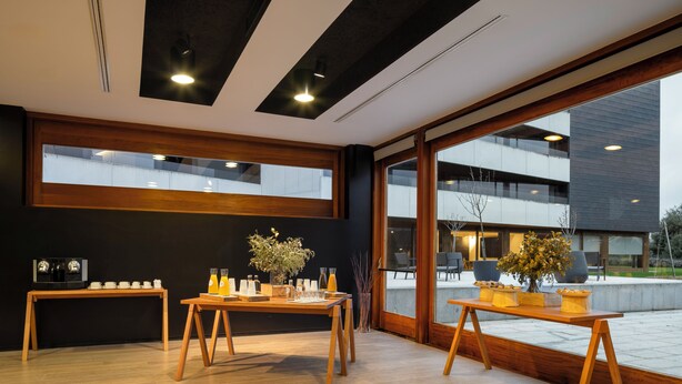 Un interior moderno con mesas de madera, plantas en macetas y grandes ventanales que ofrecen vistas de un edificio cercano.