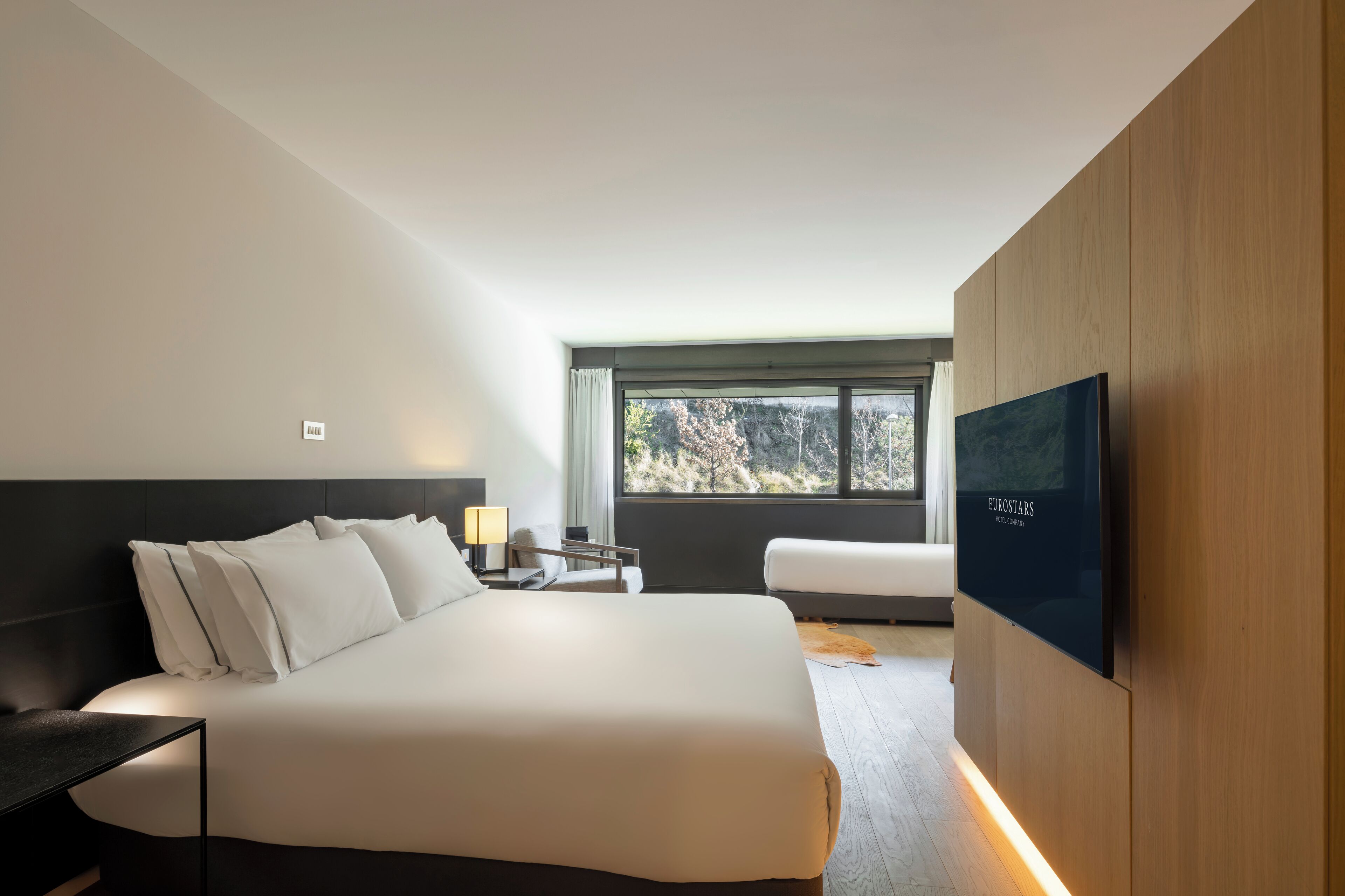 Una habitación de hotel moderna con una cama grande, televisión, mesita de noche y ventana con vista a árboles.