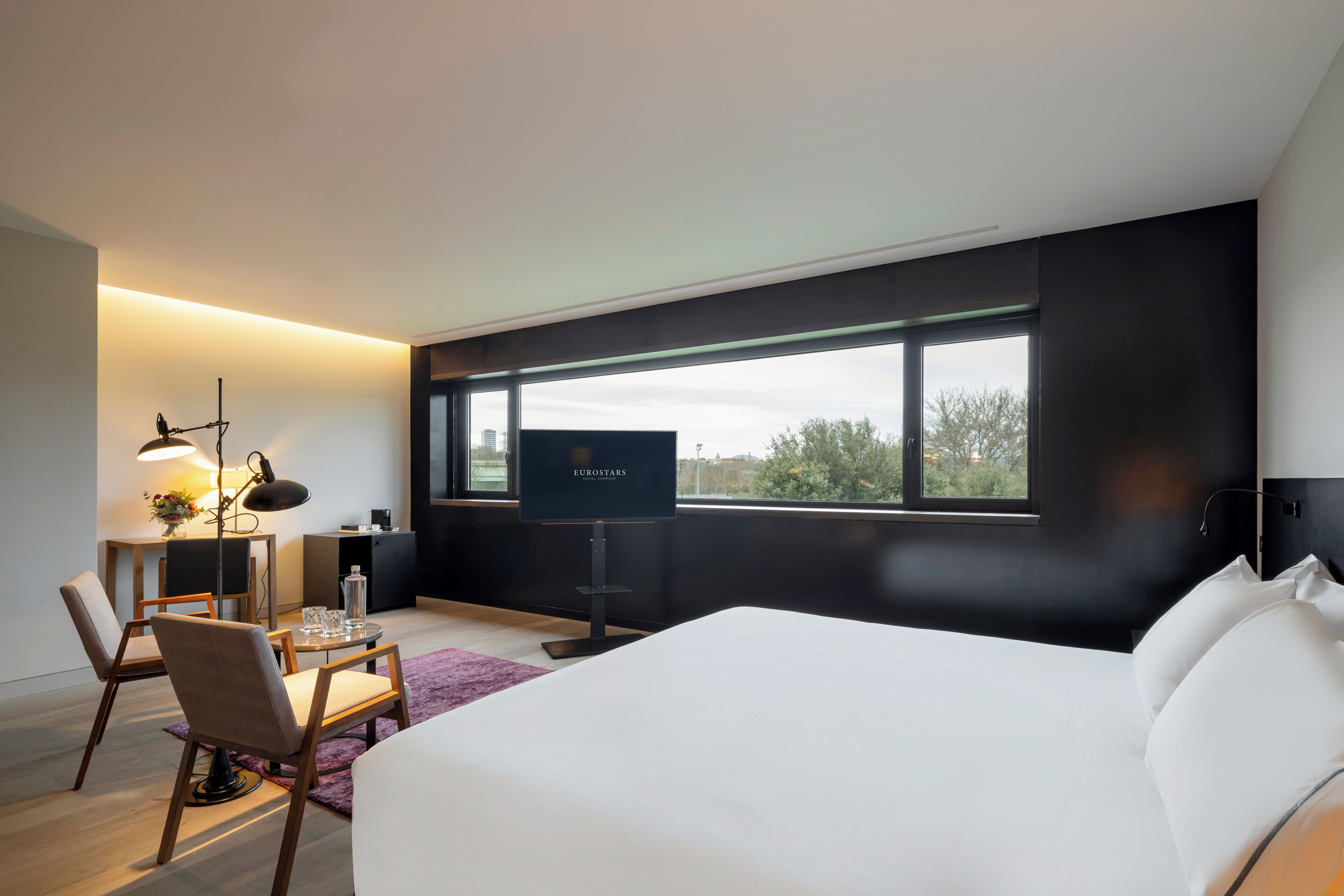 Ein modernes Hotelzimmer mit einem großen Bett, einem Schreibtisch mit Lampe, einem Fernseher und Blick ins Freie durch ein großes Fenster.
