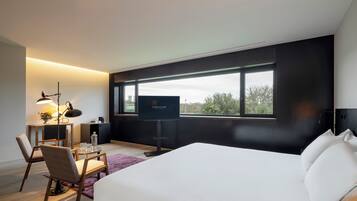 Ein modernes Hotelzimmer mit einem großen Bett, einem Schreibtisch mit Lampe, einem Fernseher und Blick ins Freie durch ein großes Fenster.