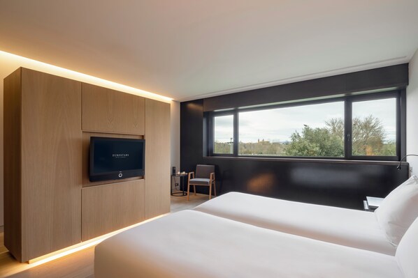 Un moderno cuarto de hotel con una cama grande, un televisor montado en un mueble de madera, un pequeño escritorio con una silla, y un ventanal que ofrece vistas al exterior.