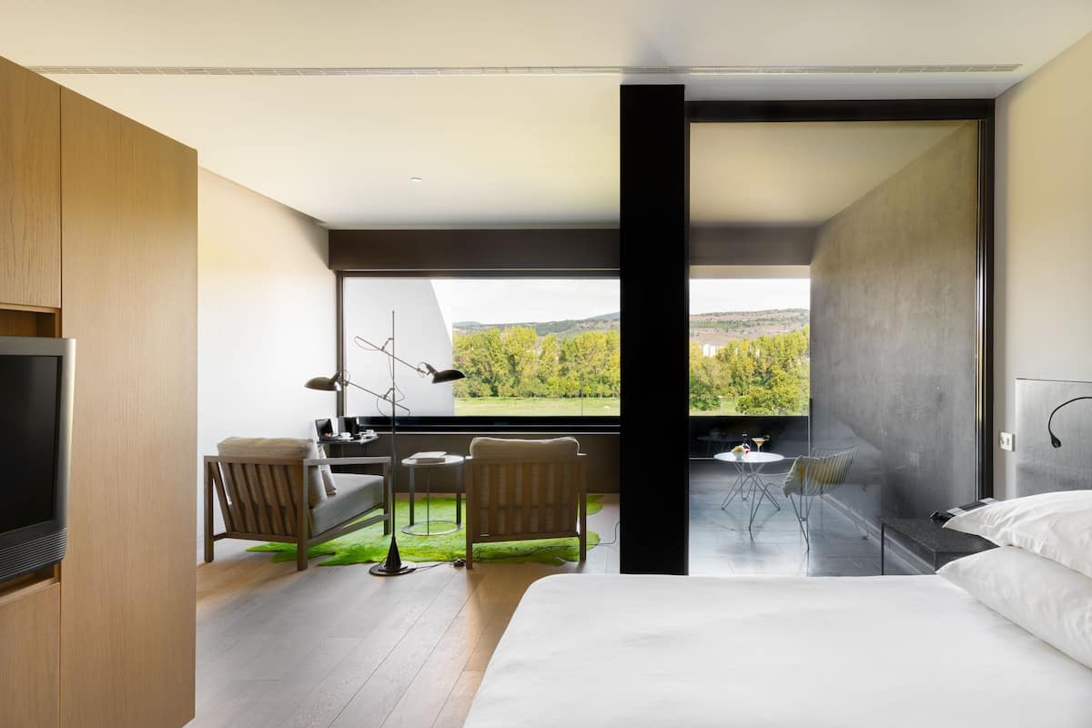 Een moderne hotelkamer met een bed, een bureau met stoel, een kleine tafel met stoel en uitzicht op een groen landschap door grote ramen.