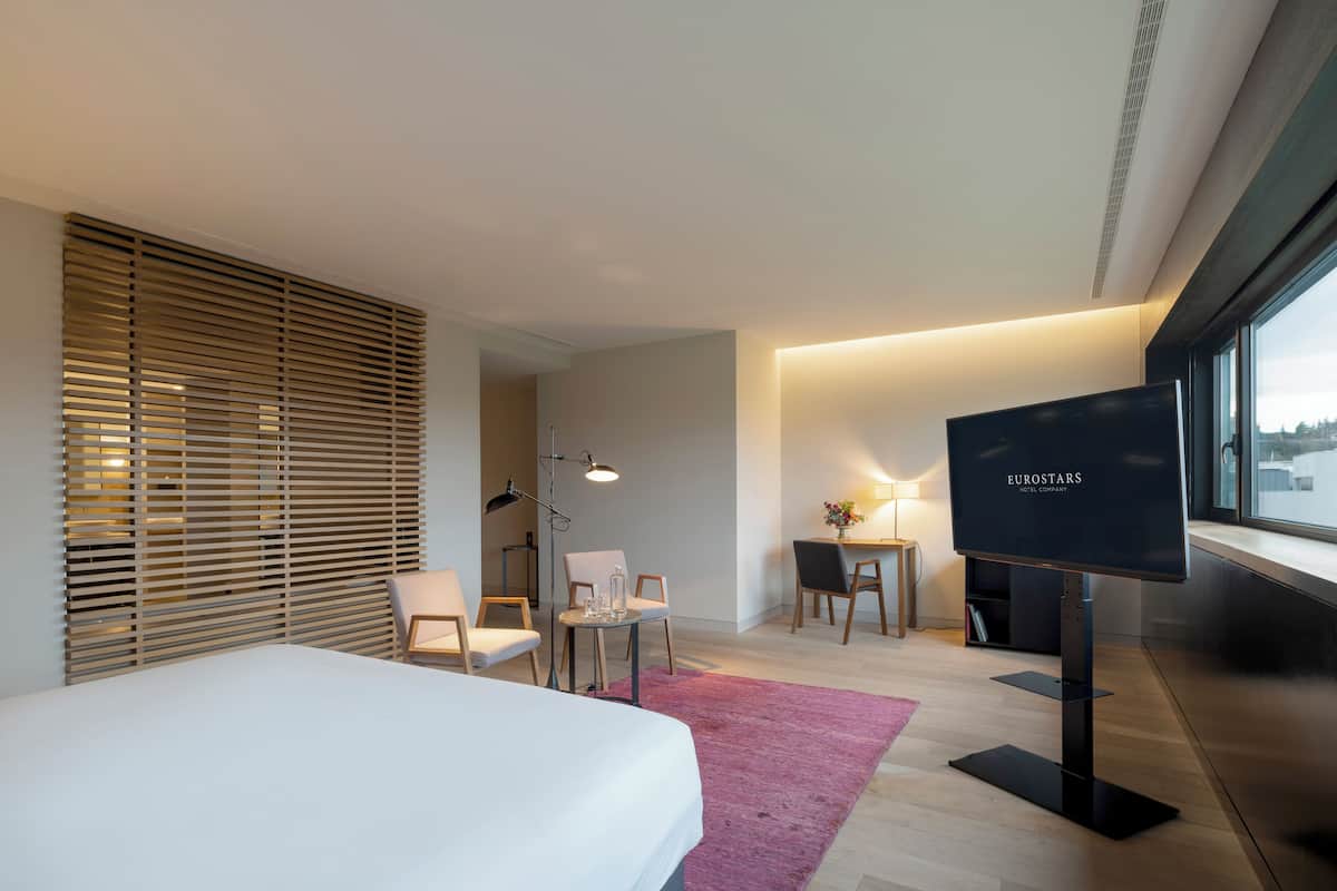 Een moderne hotelkamer met een bed, een bureau, een stoel, een televisie en een raam met zonwering.