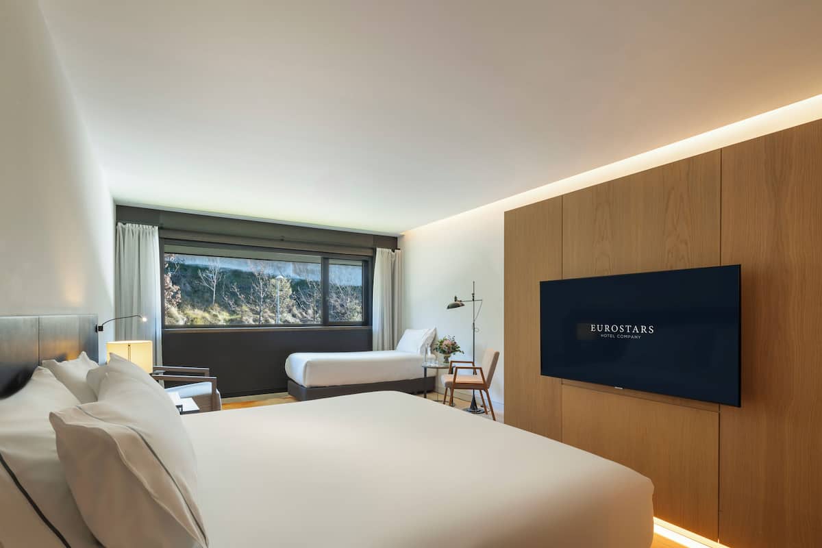 Een moderne hotelkamer met een groot bed, een bureau, een stoel, een televisie en een raam met uitzicht op bomen.