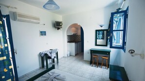 A room with a bed, a desk, a chair, a small table, and a kitchenette in the background. - Lianos Studios (Naxos)