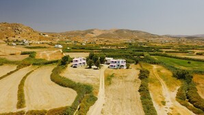 A rural landscape with two white buildings with pink roofs, surrounded by fields and a mountain range in the background. - Lianos Studios (Naxos)