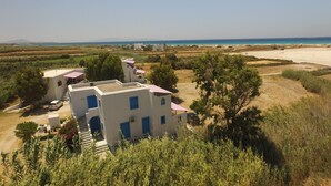 A white building with blue accents, surrounded by greenery and a beach in the background. - Lianos Studios (Naxos)