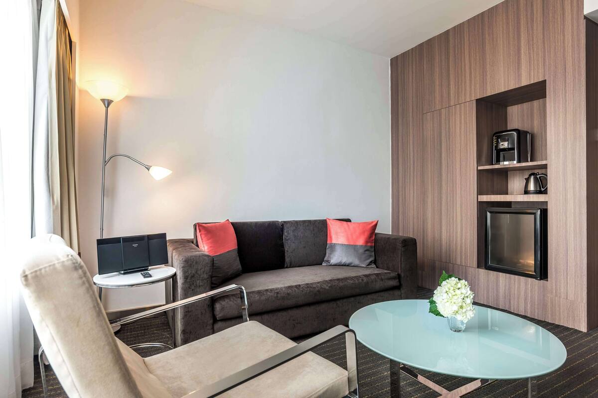 Een moderne hotelkamer met een grijze bank, een glazen salontafel, een beige fauteuil, een bijzettafeltje met een lamp en een tv aan de muur.