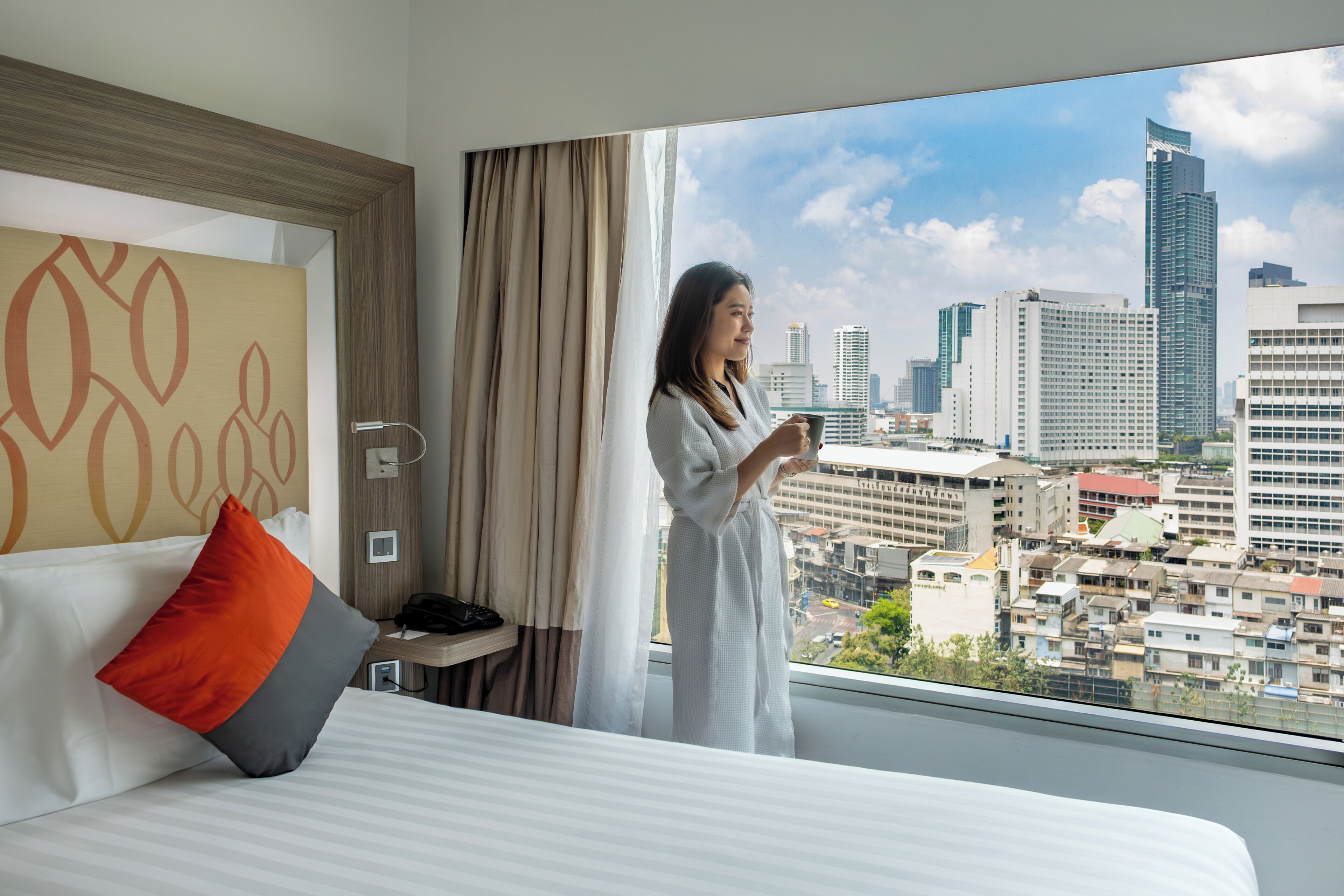 Habitación de hotel con un ventanal grande, una cama con ropa de cama blanca, almohada roja y gris, y vista panorámica de la ciudad.