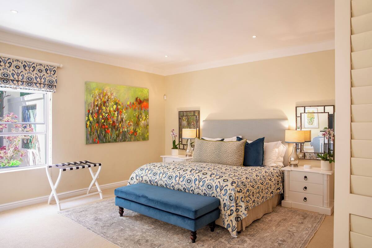 A bedroom with a bed, bedside tables, a bench, a painting, and a window.