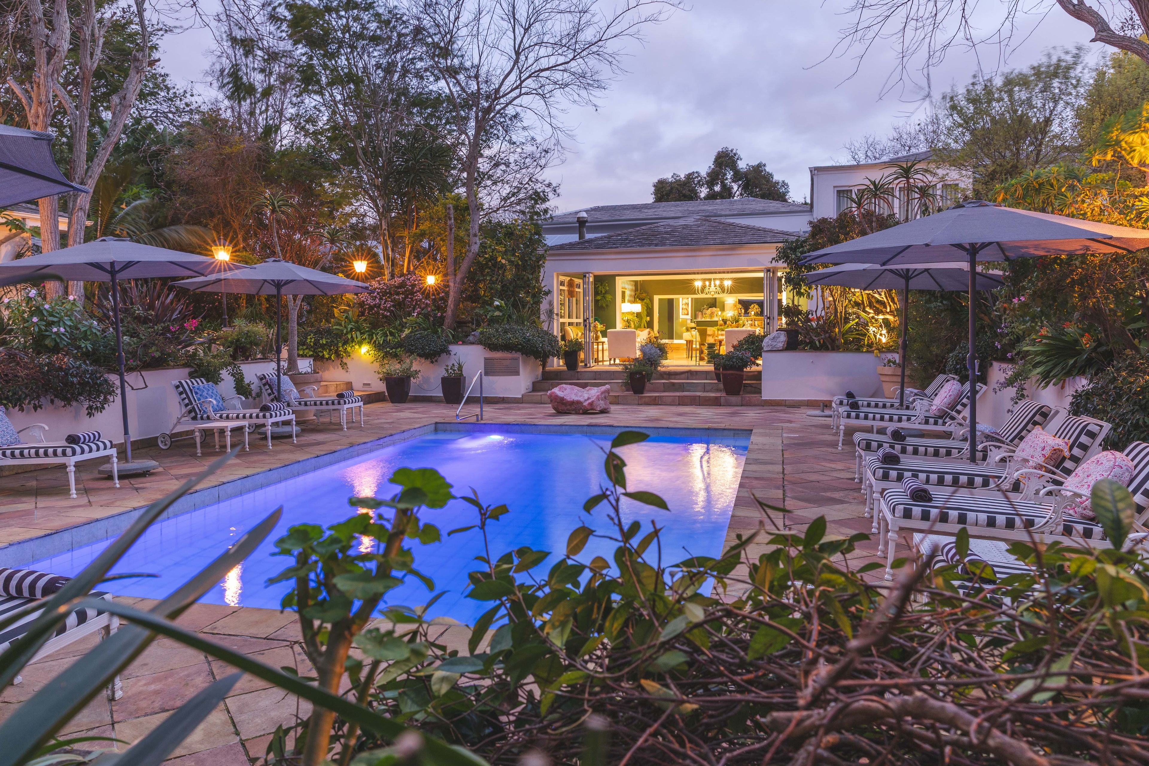 A pool area with lounge chairs and umbrellas, surrounded by lush greenery and a building with a patio.