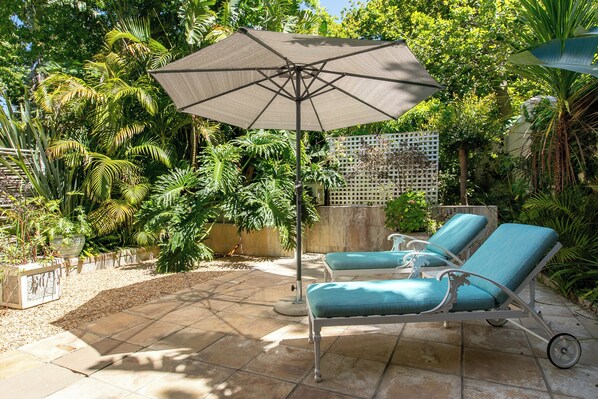 A patio with two lounge chairs and a large umbrella, surrounded by lush tropical plants.