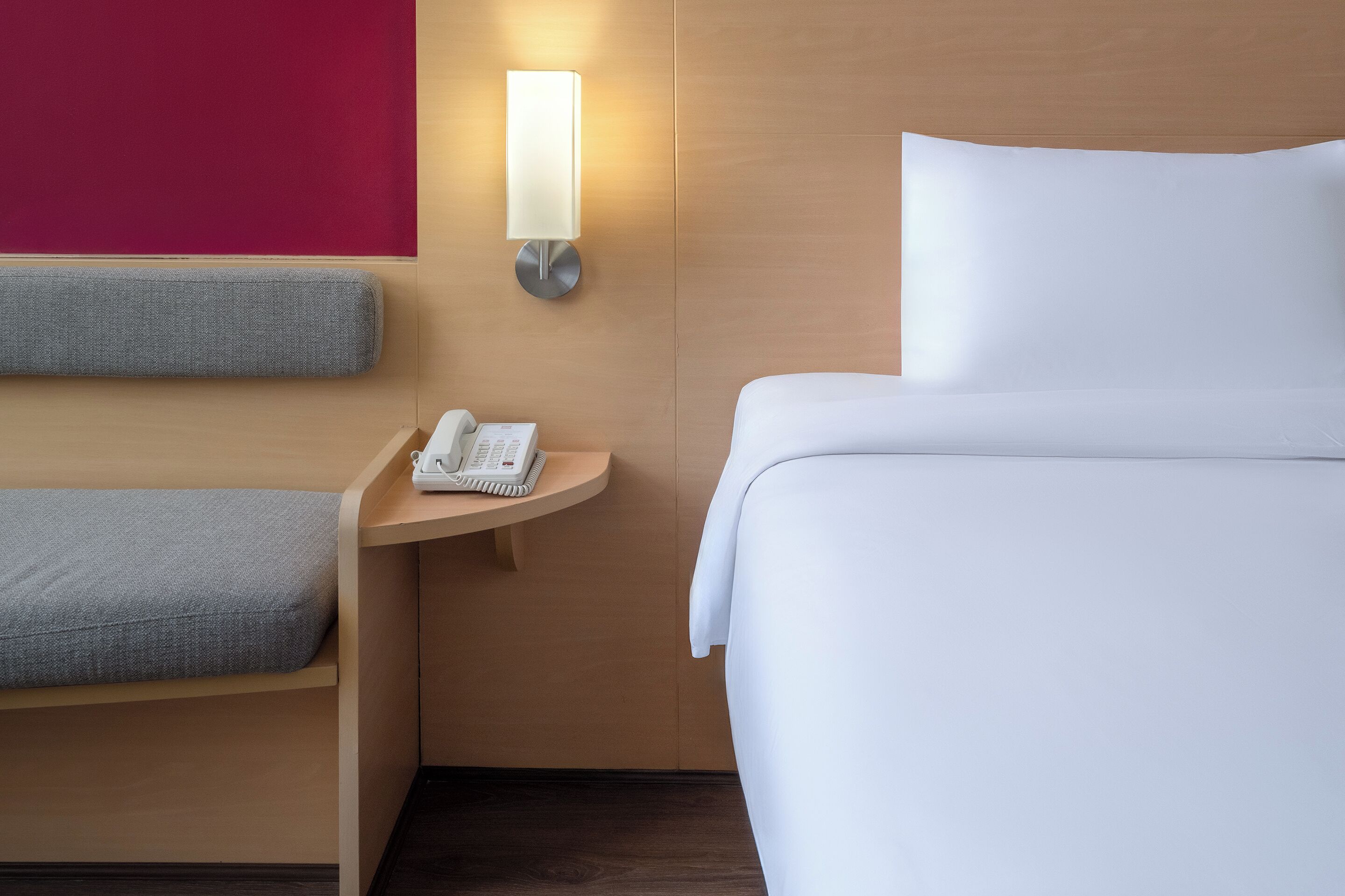 A hotel room with a bed, a bedside table with a phone, and a wall-mounted light.
